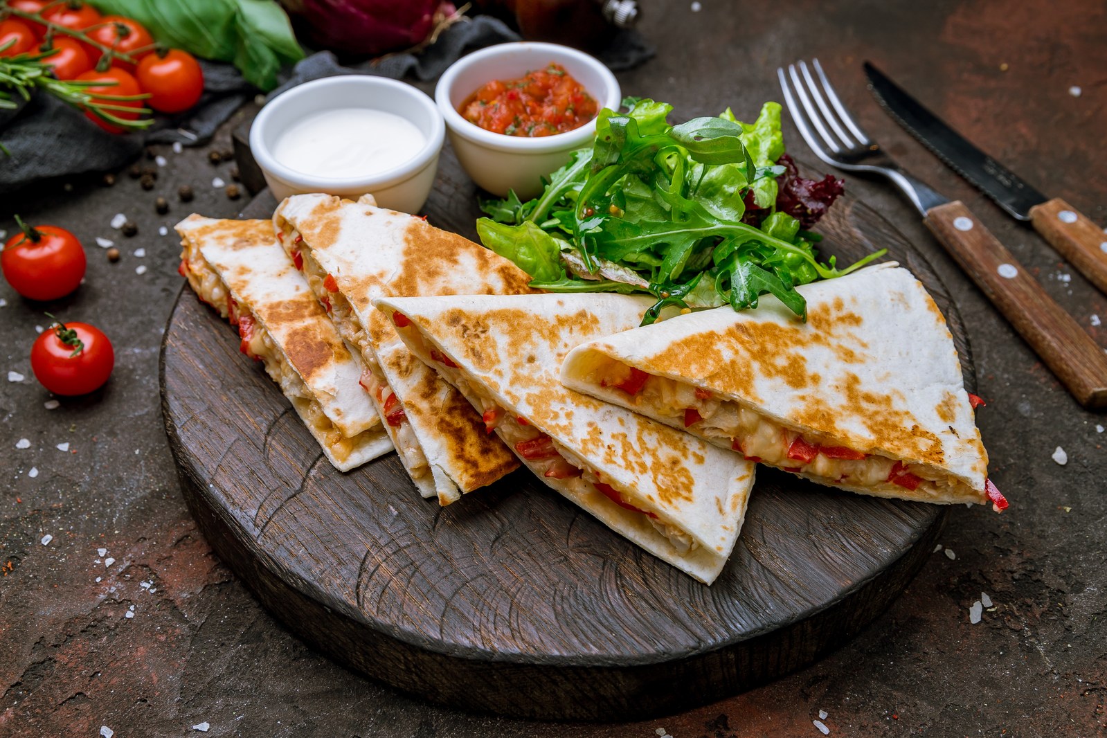 Sliced quesadillas on a wooden board with melted cheese filling, served with fresh greens, salsa, and sour cream