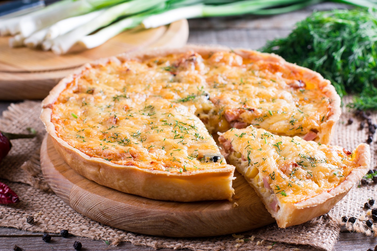 French quiche Lorraine with golden-brown Gruyère cheese top, flaky buttery crust, one slice removed showing custardy egg filling with bacon and onions on wooden board