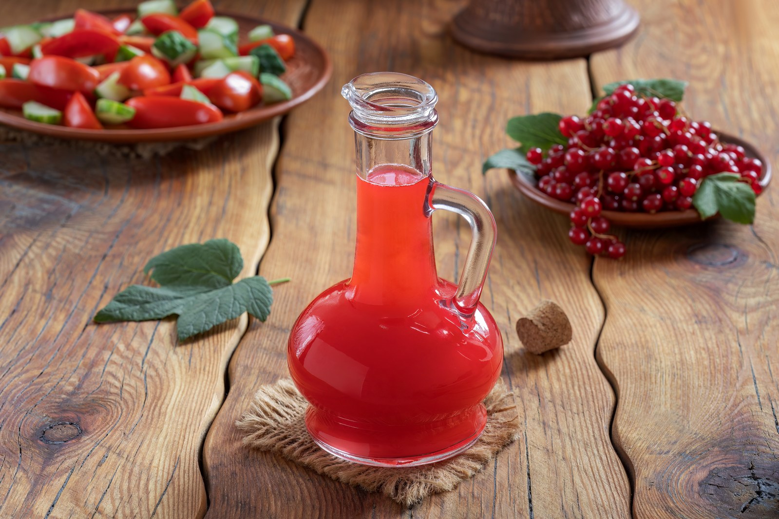 A glass cruet with handle filled with deep red vinaigrette on a wooden table with fresh vegetables and herbs in the background