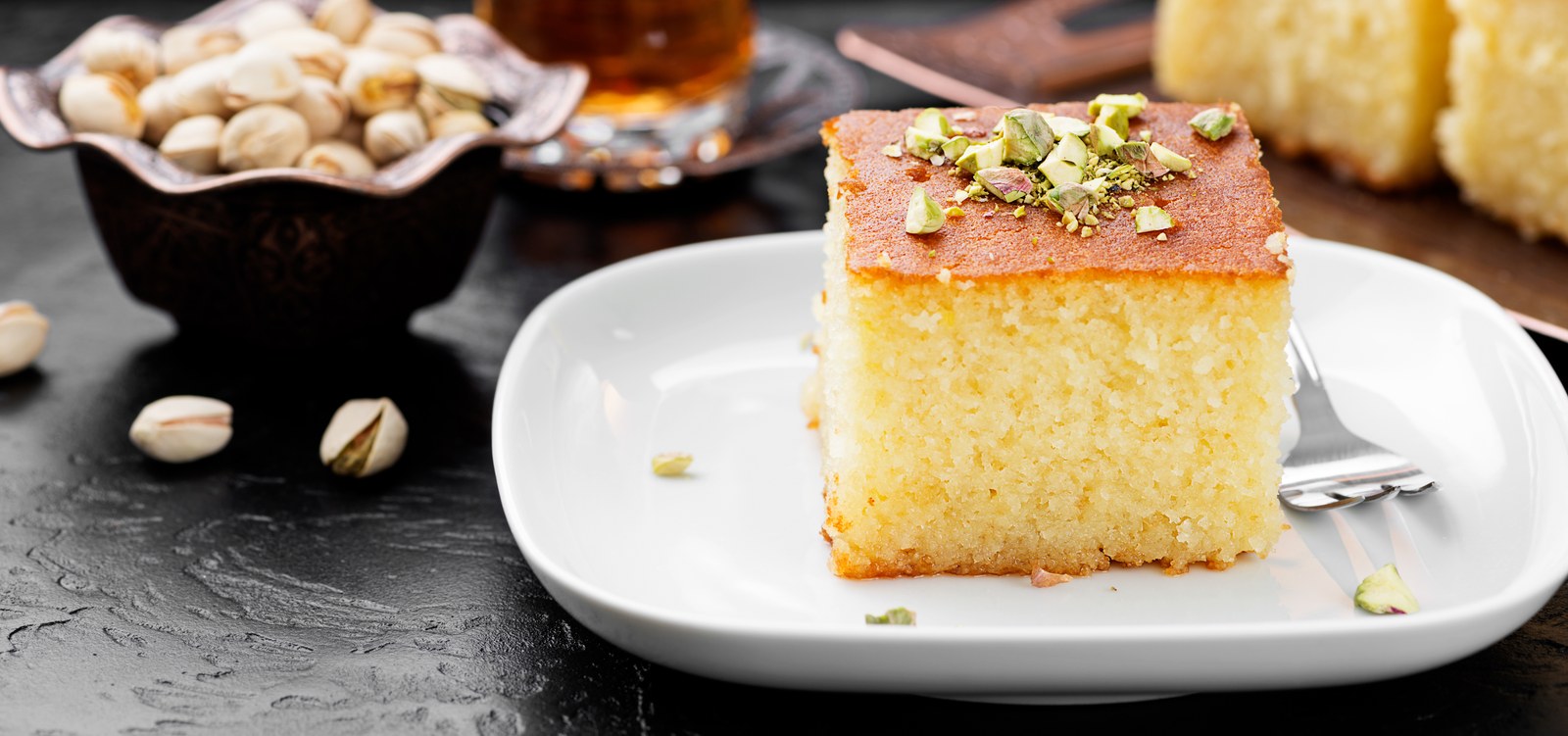 Square slice of Greek revani semolina cake topped with chopped pistachios on a white platev
