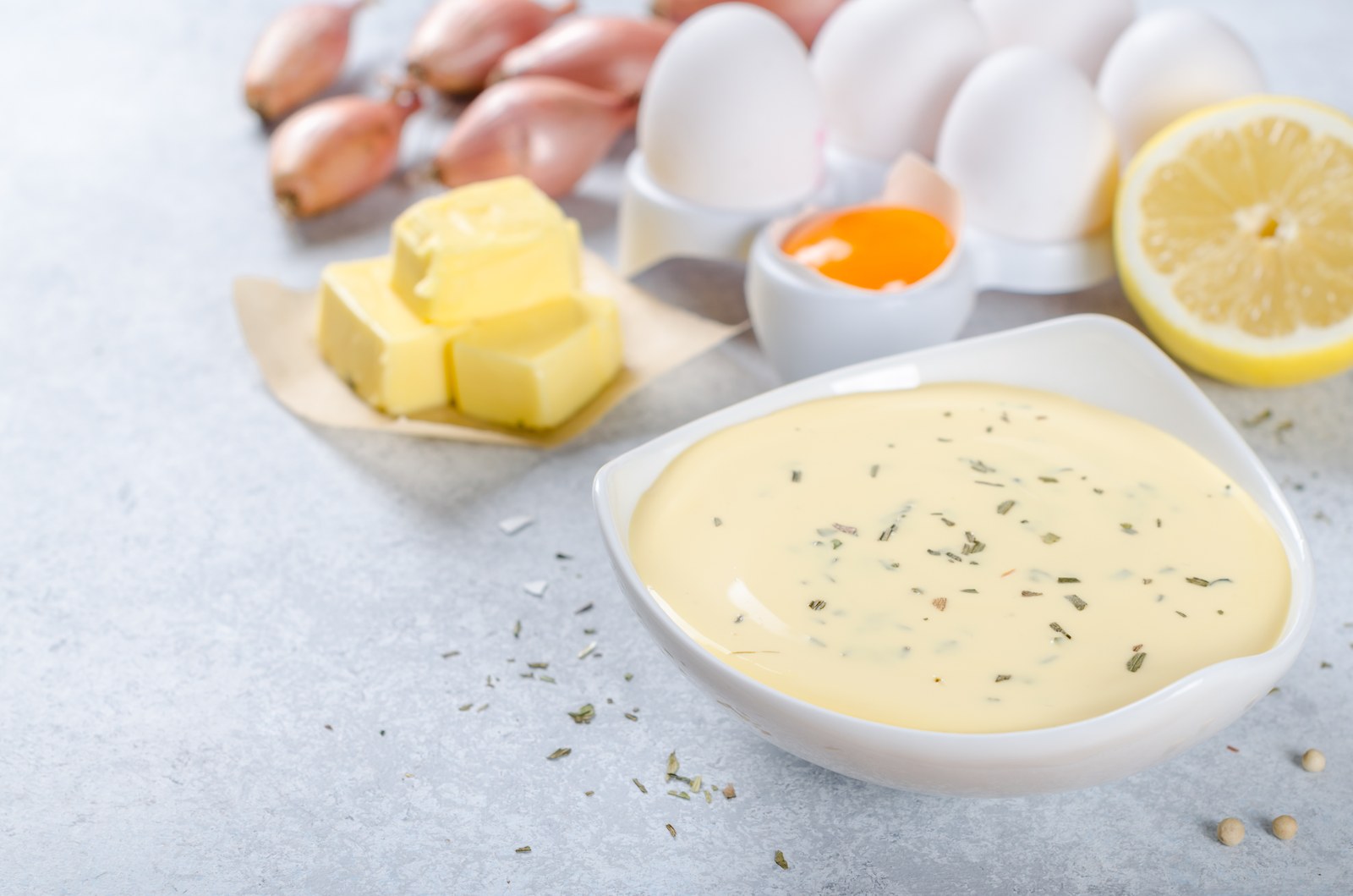 White bowl of creamy béarnaise sauce with fresh tarragon, surrounded by ingredients including butter, eggs, shallots, lemon, and peppercorns