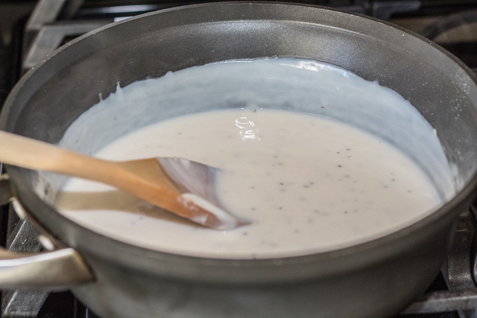 Creamy white sausage gravy with black pepper specks being stirred with a wooden spoon in a gray skillet on a stovetop