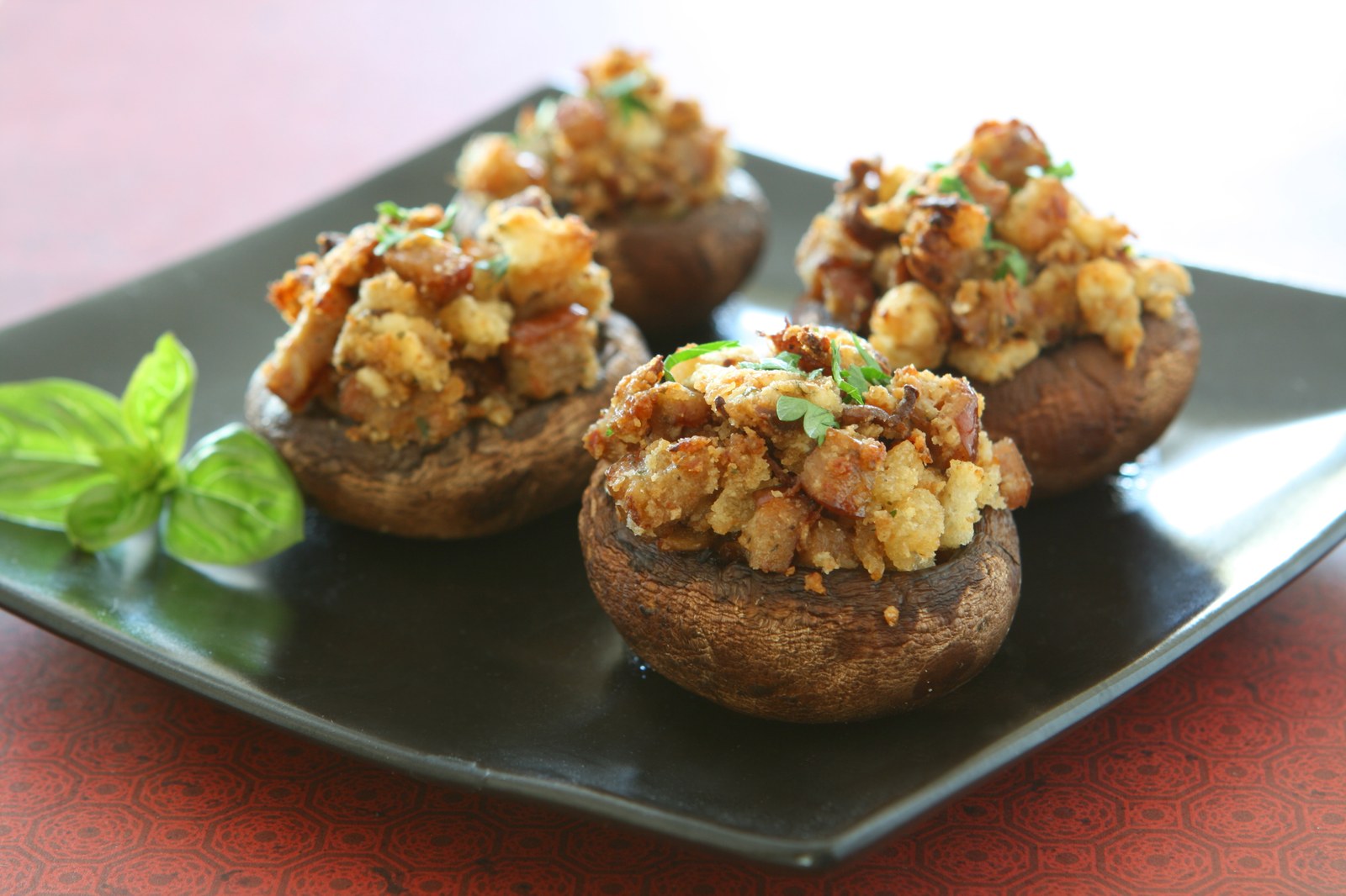 Four large brown mushroom caps filled with golden sausage and breadcrumb stuffing, garnished with fresh parsley, arranged on a black rectangular plate