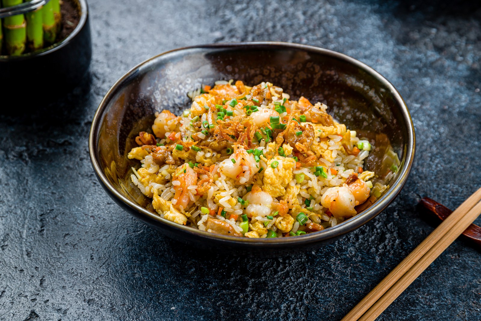 Fried rice loaded with shrimp, scrambled eggs, and green onions in a dark brown bowl with chopsticks beside it