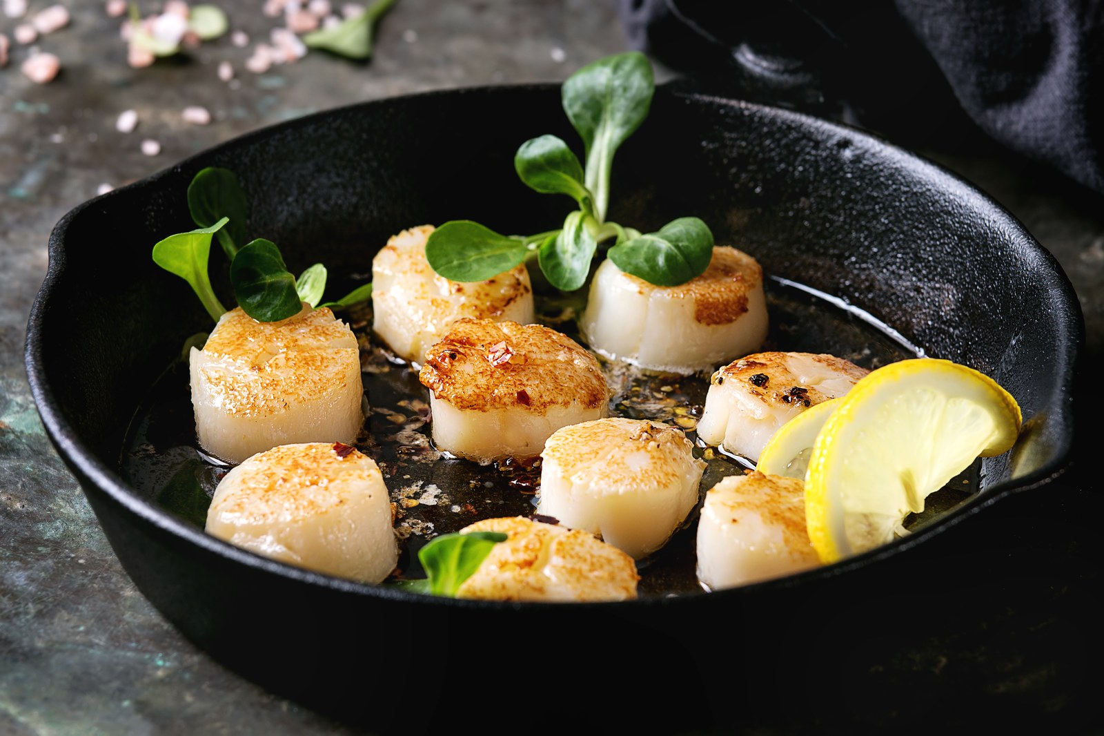 Seven golden brown sea scallops arranged in a black cast iron pan with microgreens and a lemon wedge