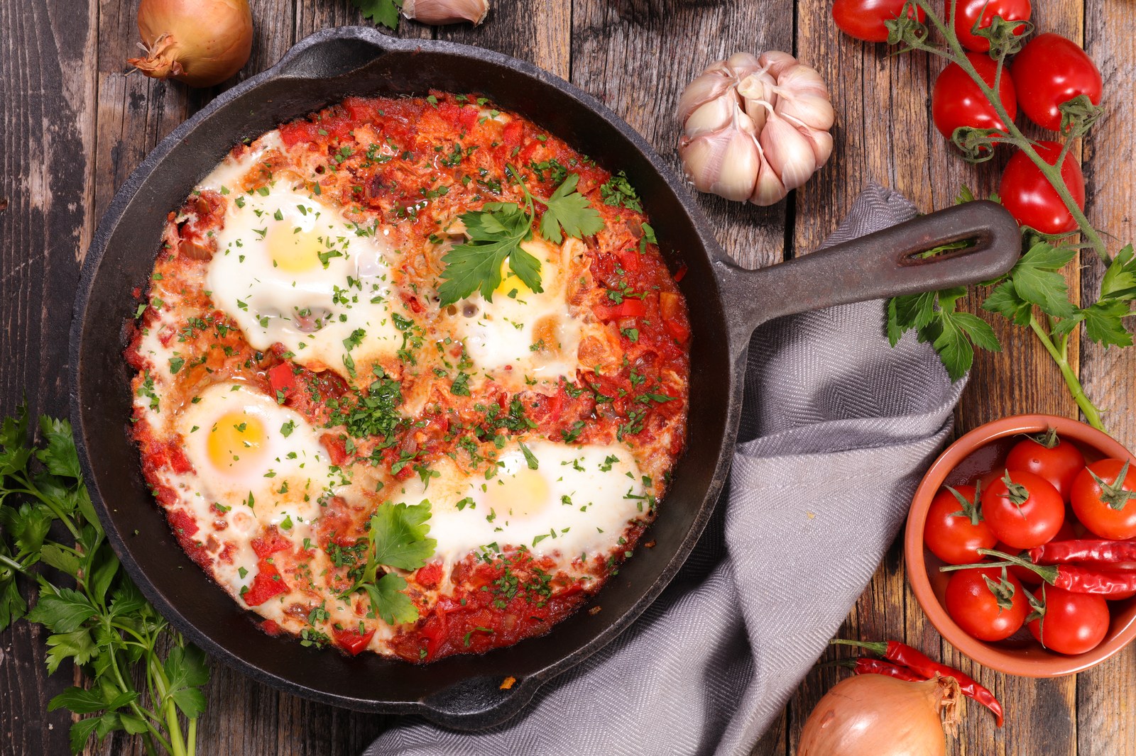 Cast iron skillet of shakshuka with eggs poached in spiced tomato sauce, garnished with fresh parsley and surrounded by raw ingredients