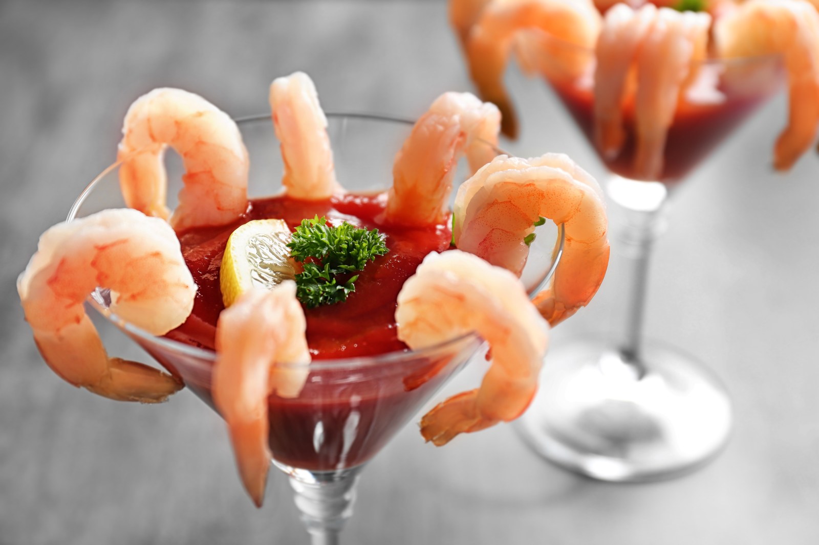 Five large cooked shrimp arranged around the rim of a cocktail glass filled with red cocktail sauce, garnished with parsley and lemon