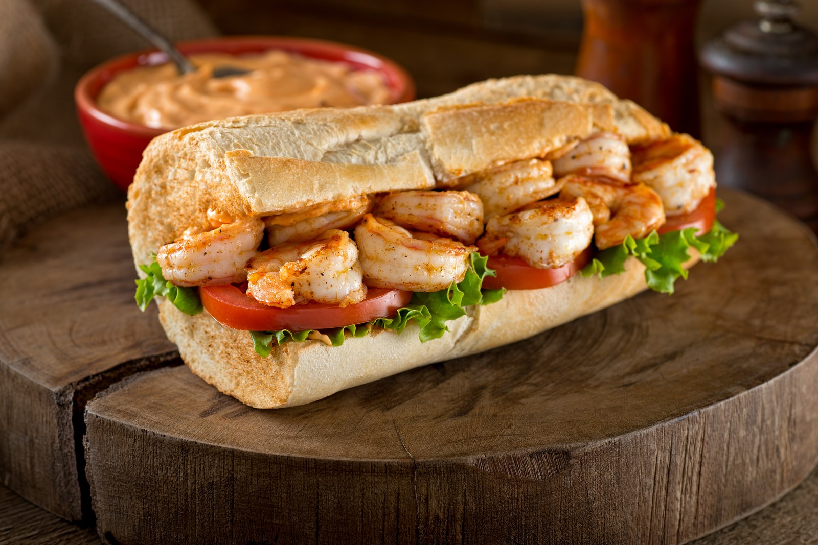 A New Orleans style po'boy sandwich on a wooden board filled with fried shrimp, lettuce, and tomato slices on French bread with remoulade sauce in background