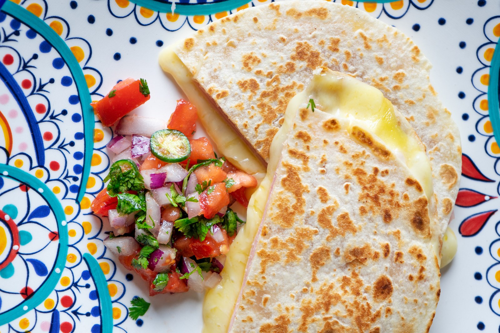 Two golden grilled flour tortillas filled with melted cheese on a colorful Mexican ceramic plate, served with fresh pico de gallo on the side