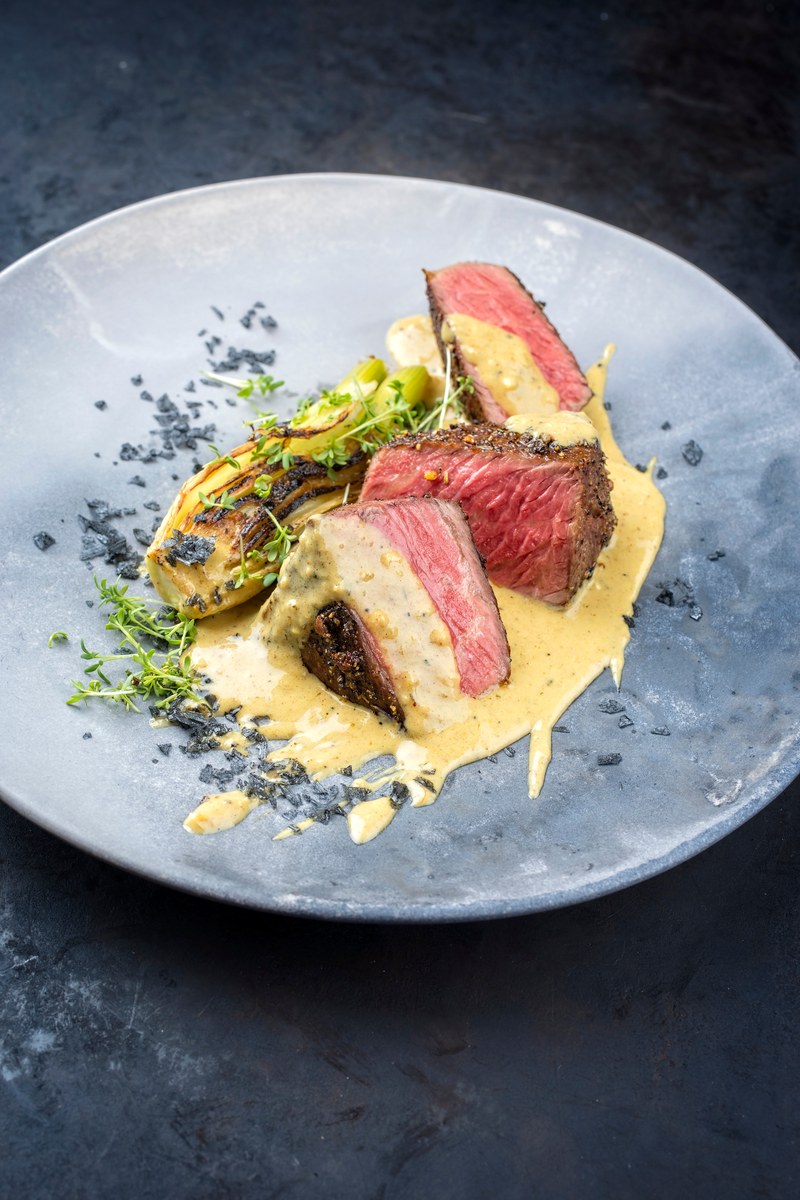 Sliced medium rare sirloin steak with pink center arranged on a blue plate, covered in creamy peppercorn sauce and garnished with microgreens and caramelized onions