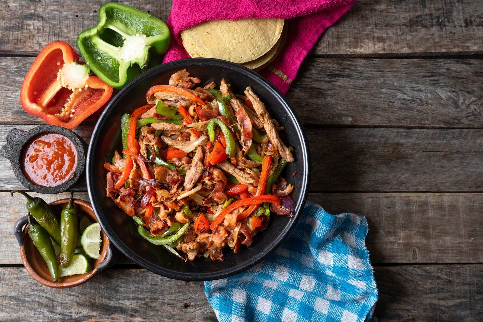 Black cast iron skillet filled with strips of grilled skirt steak, red and green bell peppers, and caramelized onions, surrounded by fresh peppers, salsa, lime wedges, and corn tortillas