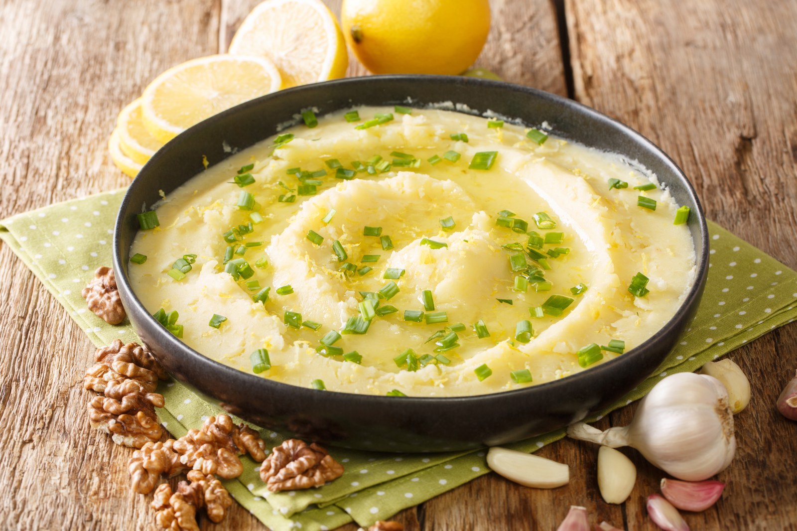Dark bowl filled with smooth white garlic potato dip drizzled with olive oil and topped with fresh chives, with lemon slices, garlic cloves, and walnuts arranged around it
