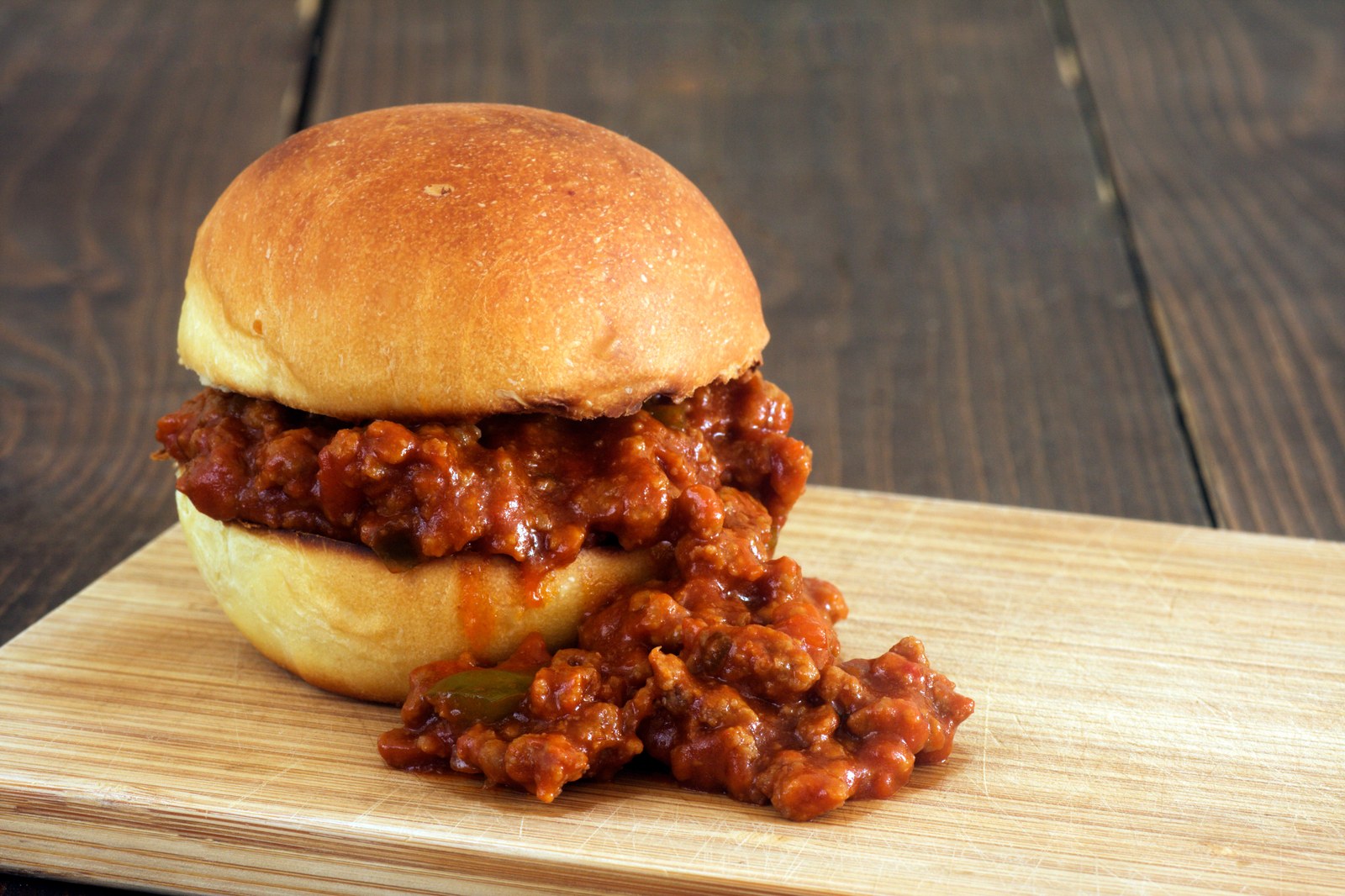 A soft hamburger bun on a wooden board filled with saucy ground beef mixture in tomato sauce, with extra filling spilling onto the board