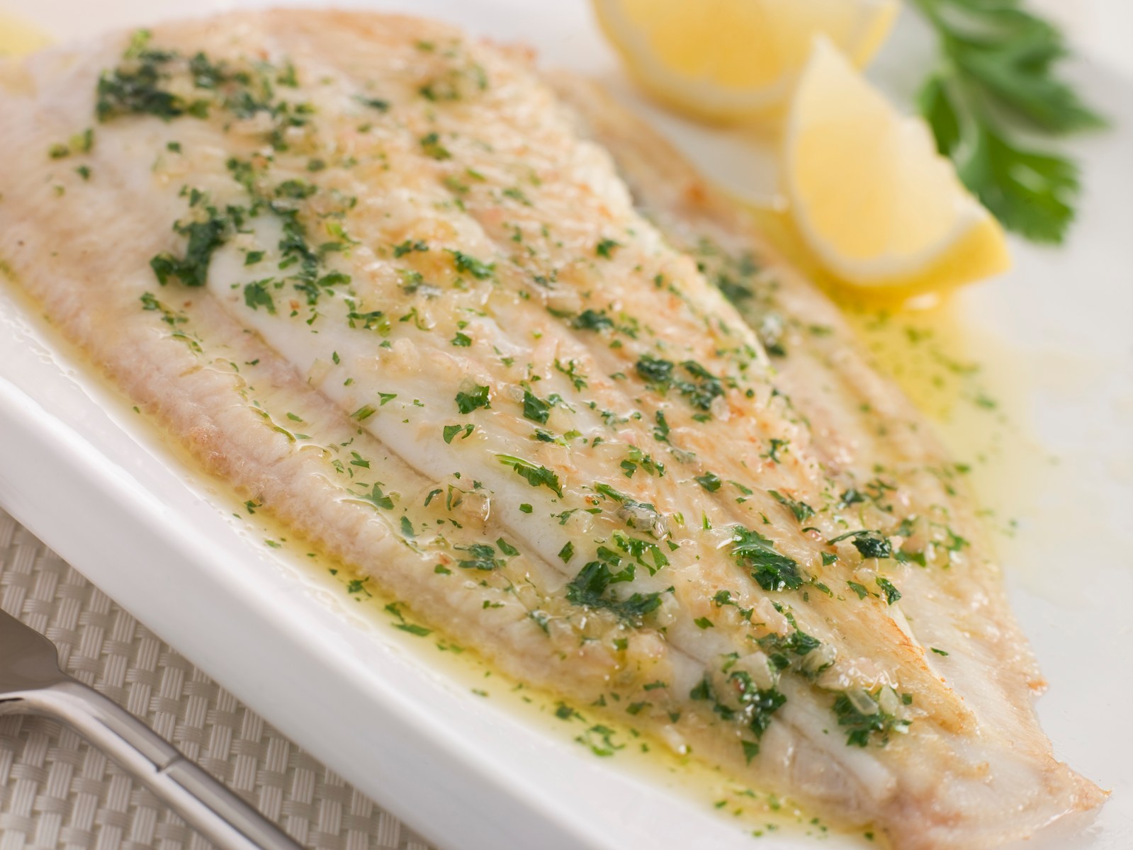 Pan fried sole fillet topped with brown butter sauce, lemon, and chopped parsley on a white plate