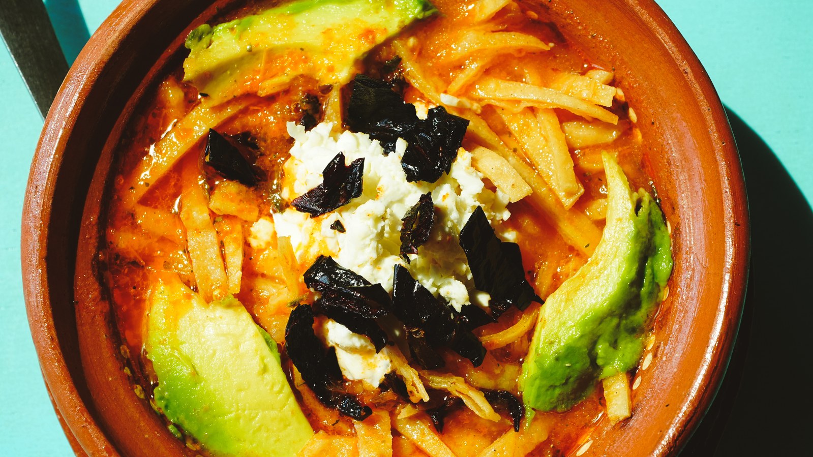 Mexican tortilla soup in a rustic terracotta bowl topped with crispy tortilla strips, avocado slices, crumbled white cheese, and dark pasilla chile pieces in red broth