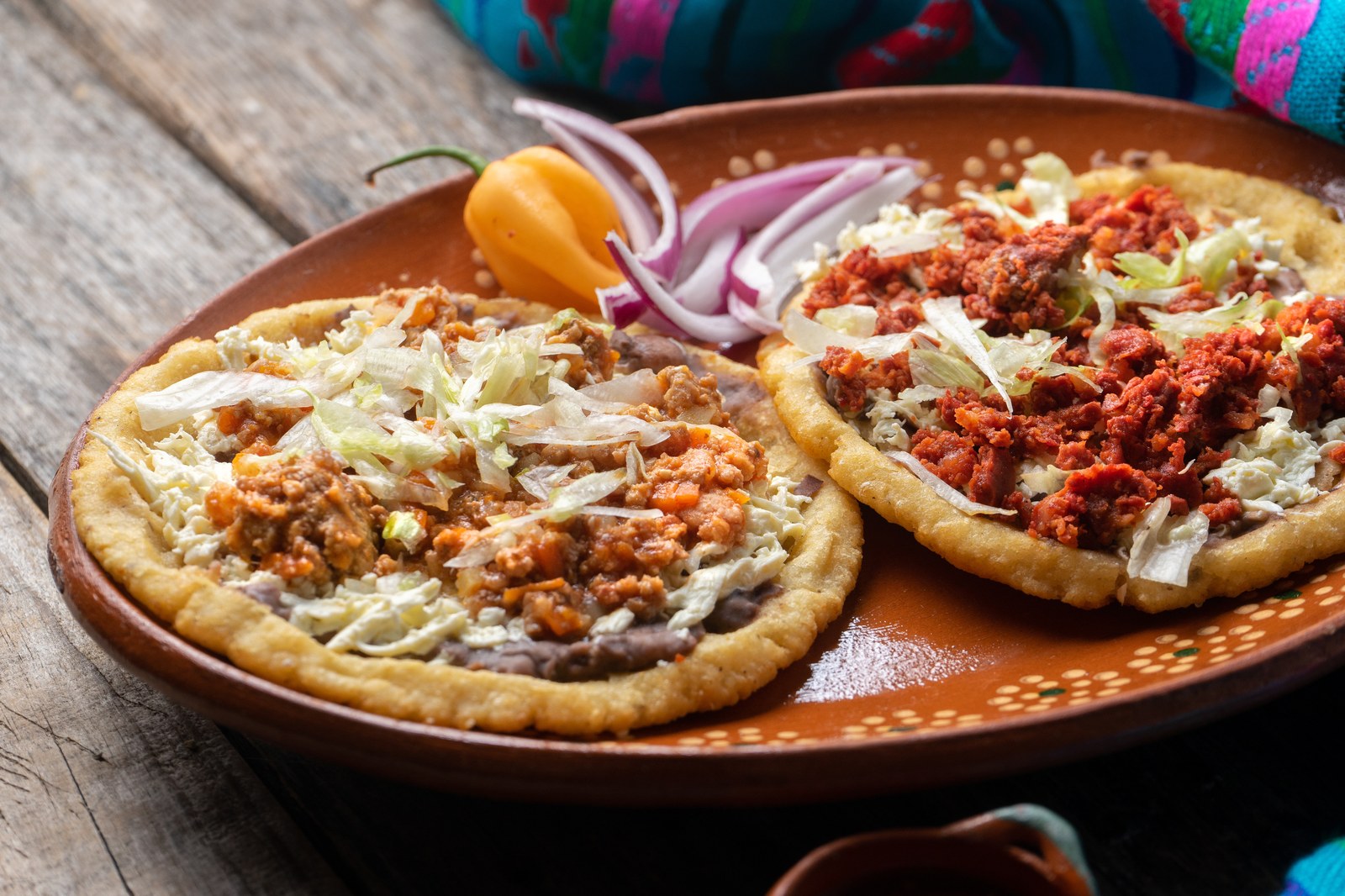 Two thick handmade corn masa bases with pinched edges on a traditional clay plate, one topped with regular seasoned beef and one with red chorizo, both garnished with refried beans, shredded lettuce, and sliced purple onions