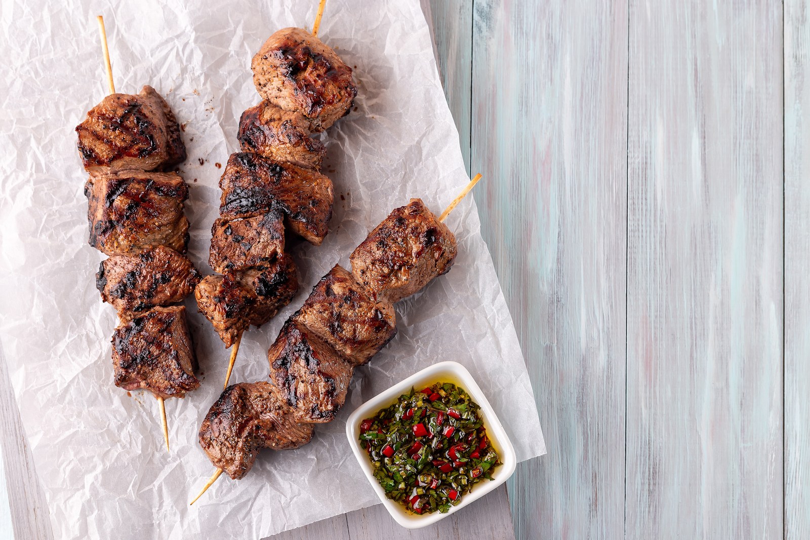 Grilled beef skewers on wooden sticks with green chimichurri sauce