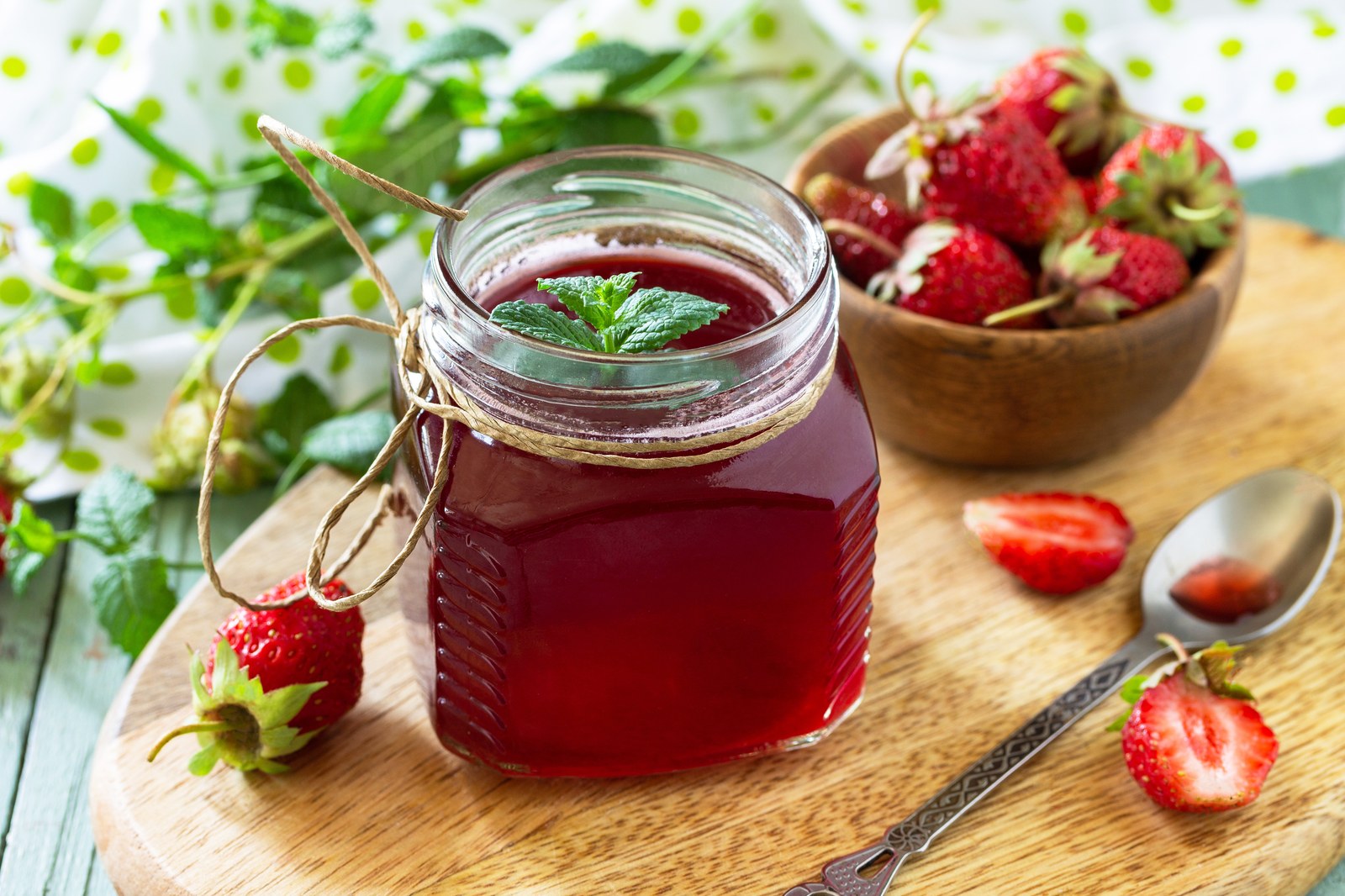 Homemade strawberry syrup in a clear glass jar with twine tied around the rim, garnished with fresh mint leaf, served on a wooden cutting board with a bowl of fresh ripe strawberries