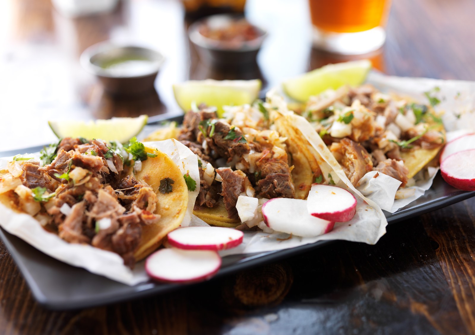 Mexican street tacos on griddled corn tortillas filled with shredded braised beef, topped with diced white onions, fresh cilantro, crumbled queso fresco, and sliced radishes on a dark wooden serving board