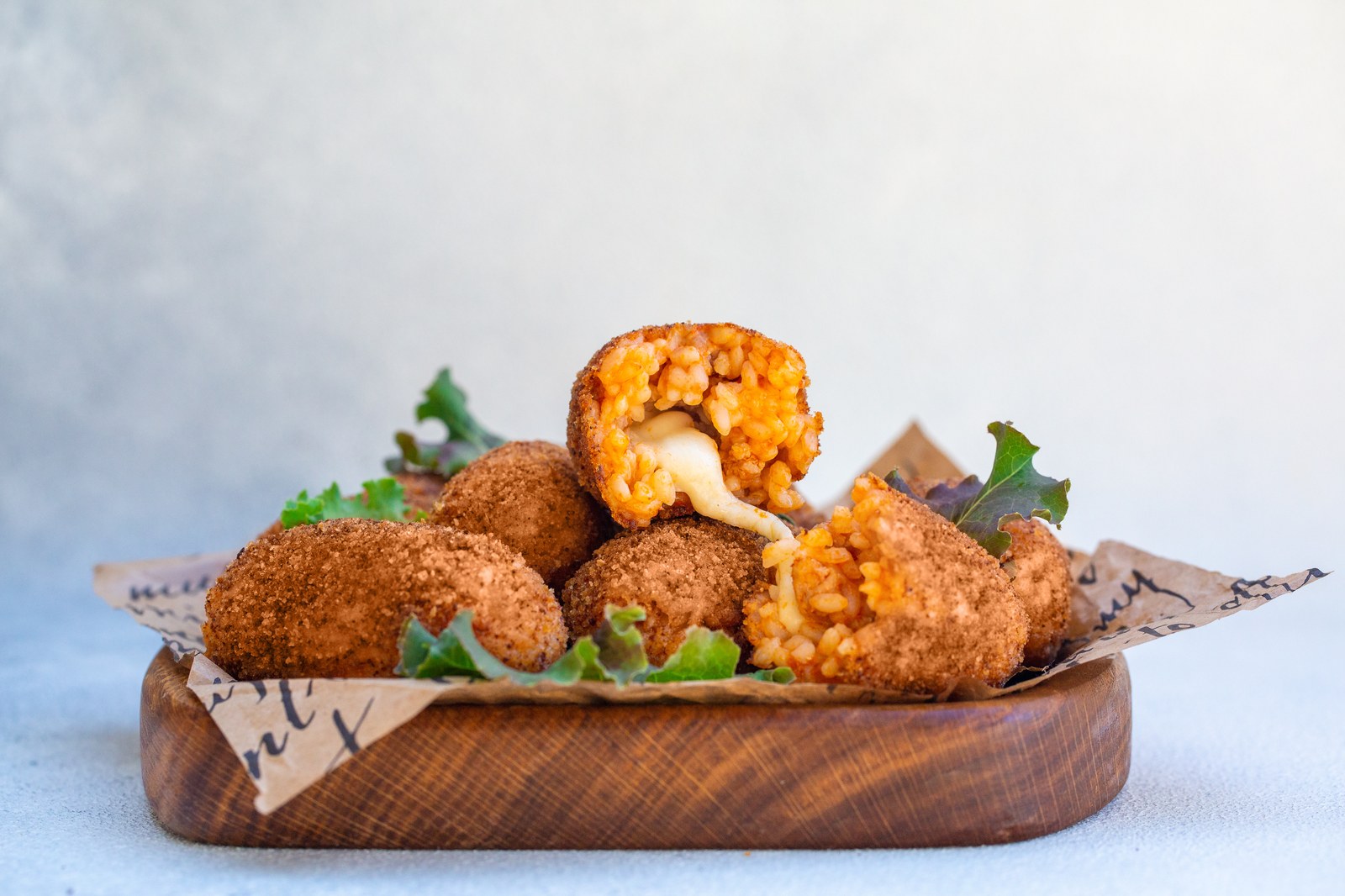 Roman rice croquettes with golden breadcrumb coating stacked on a wooden board lined with parchment paper, one cut open revealing creamy saffron risotto with stretching melted mozzarella cheese center