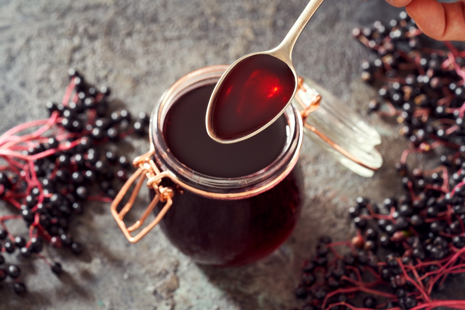 Deep burgundy elderberry syrup in glass clip-top jar, thick glossy consistency dripping from gold spoon, fresh elderberries scattered around