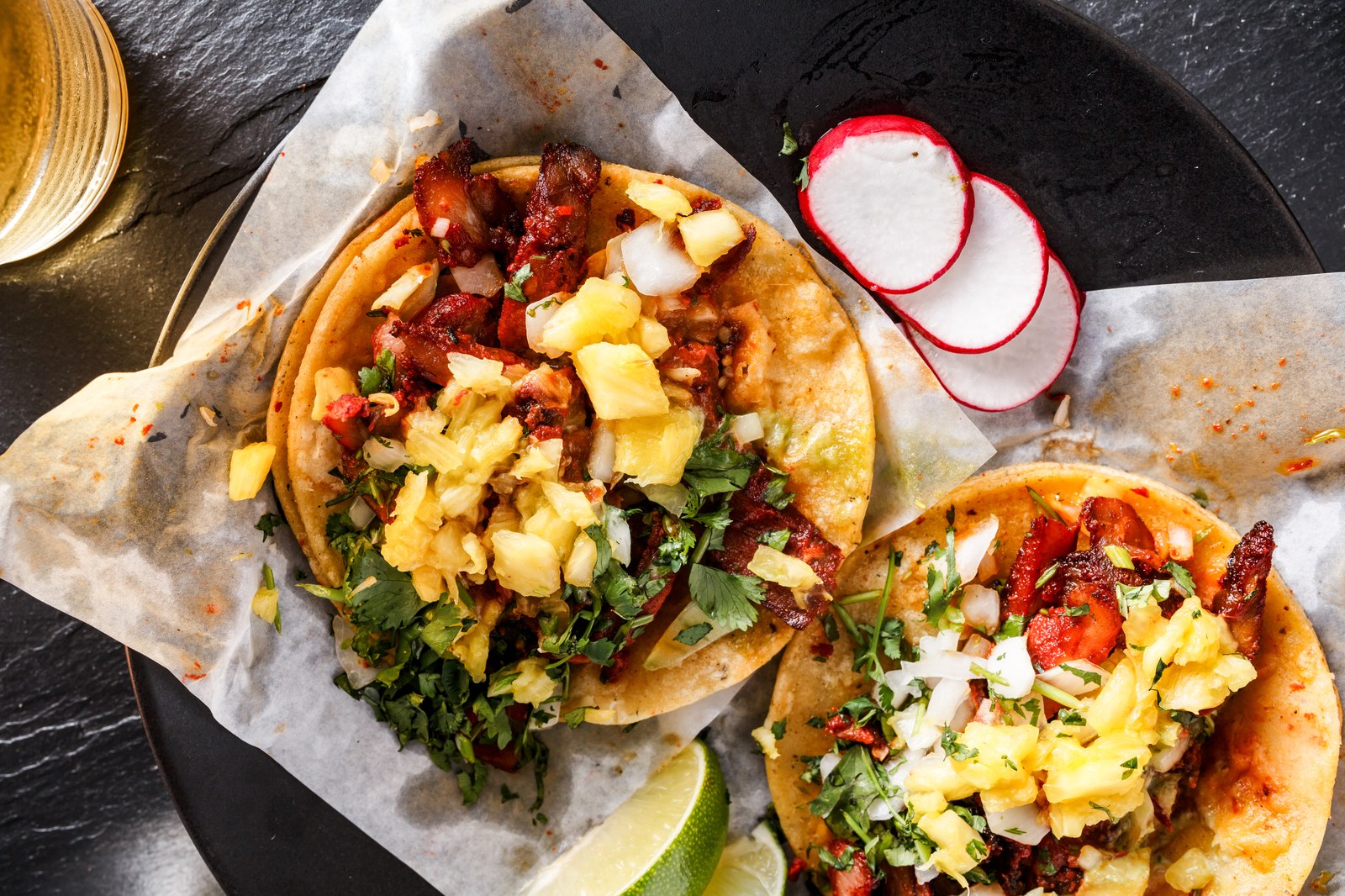 Authentic tacos al pastor on charred corn tortillas piled high with thin sliced marinated pork, chunks of grilled pineapple, diced white onions, chopped fresh cilantro, and sliced radishes on parchment paper with lime wedges