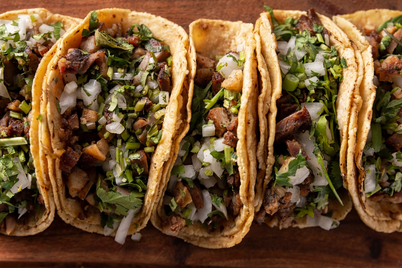 Row of traditional tacos de suadero on soft double stacked corn tortillas filled with slow braised beef brisket, topped with finely diced white onions and fresh chopped cilantro on a rustic wooden cutting board