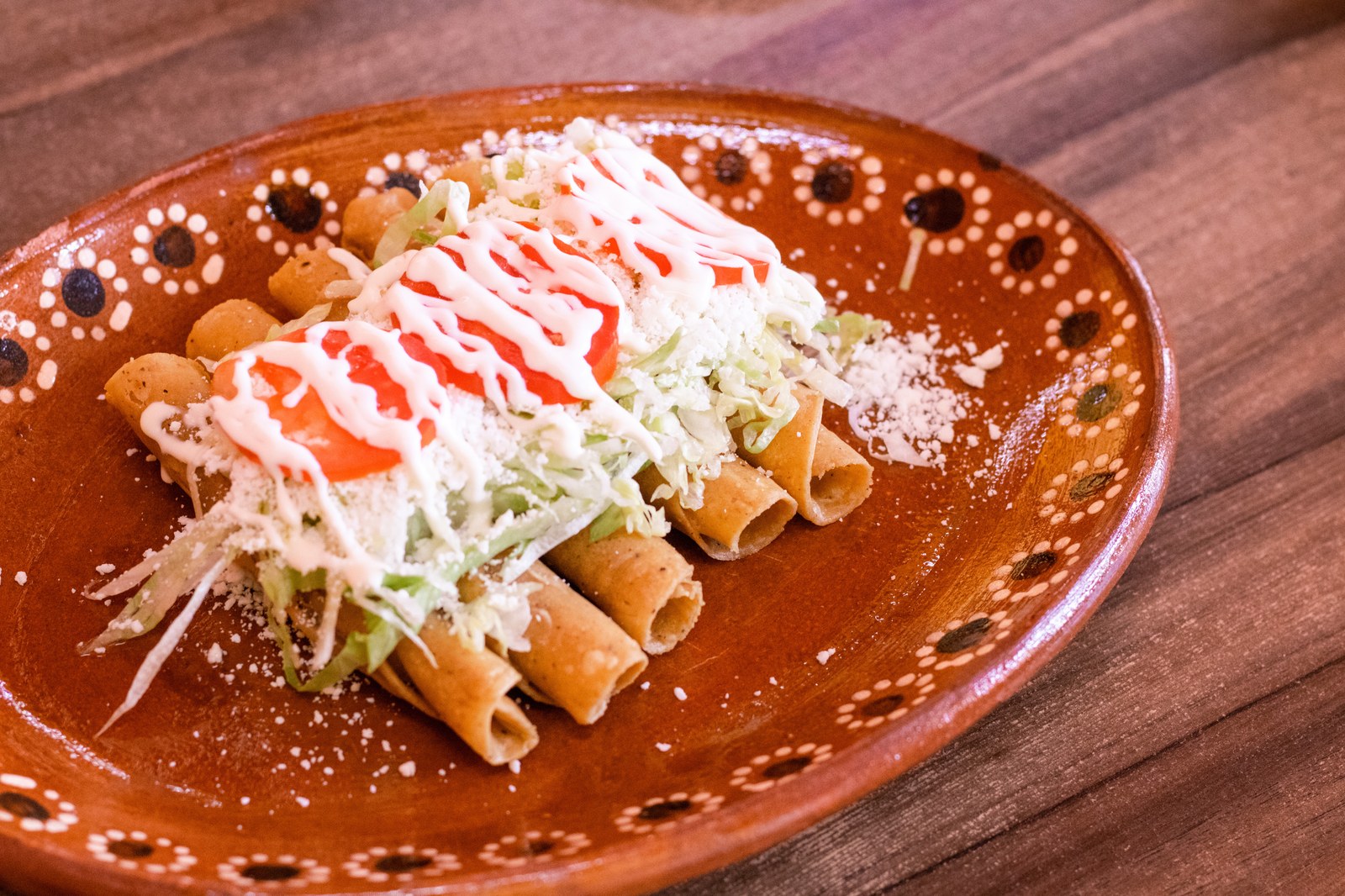 Crispy golden rolled tacos dorados on a traditional Mexican ceramic plate with hand painted decorative pattern, topped with shredded green cabbage, Mexican crema drizzle, crumbled queso fresco, and red salsa