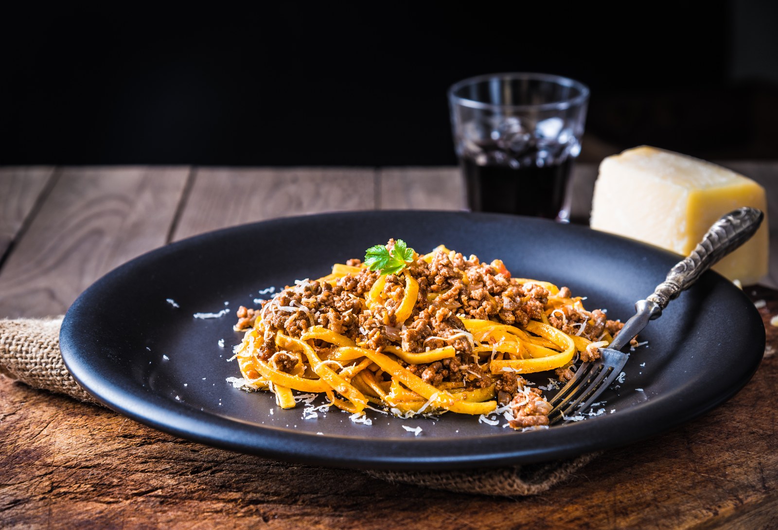 Fresh egg tagliatelle pasta ribbons tossed with rich slow simmered Bolognese meat sauce made with ground beef and tomatoes, topped with freshly grated Parmigiano Reggiano cheese and parsley garnish on a matte black plate
