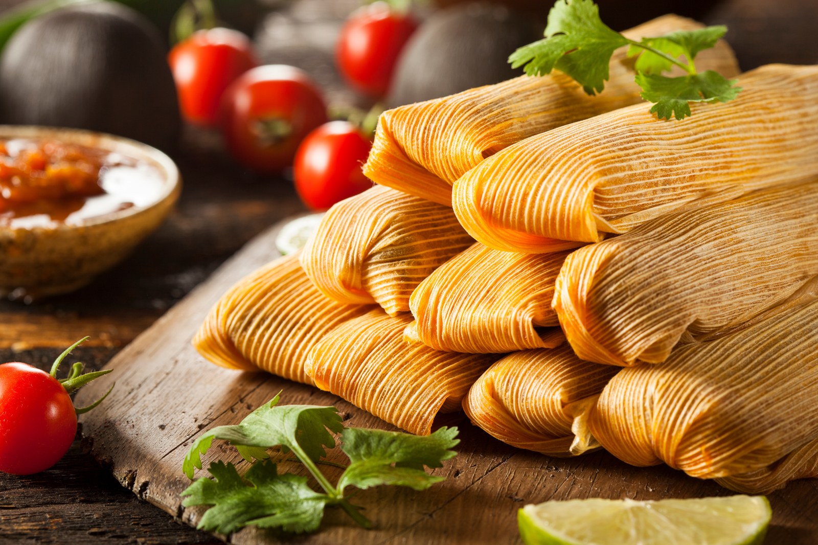 Stack of homemade tamales wrapped in dried corn husks with masa dough showing at the tops, arranged on a wooden cutting board with fresh cilantro, cherry tomatoes, and lime wedges