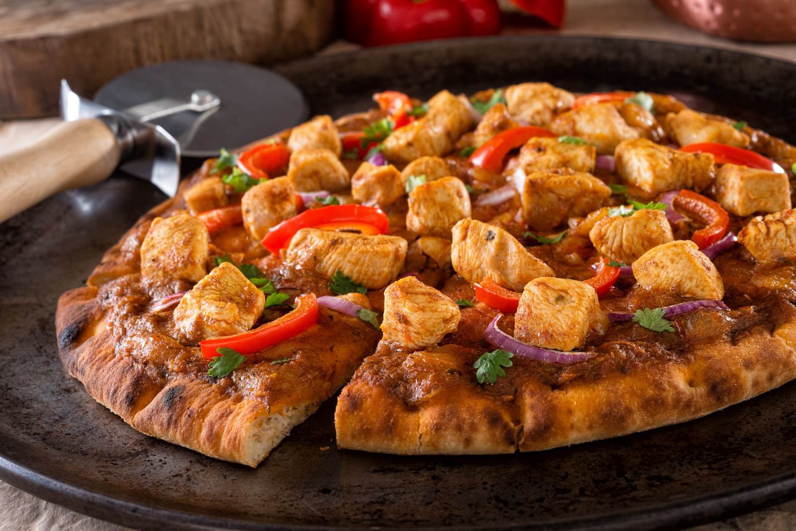 Indian fusion naan pizza topped with tandoori spiced chicken pieces, roasted red bell peppers, purple onions, and fresh cilantro on charred flatbread, sliced on a dark round pan with pizza cutter