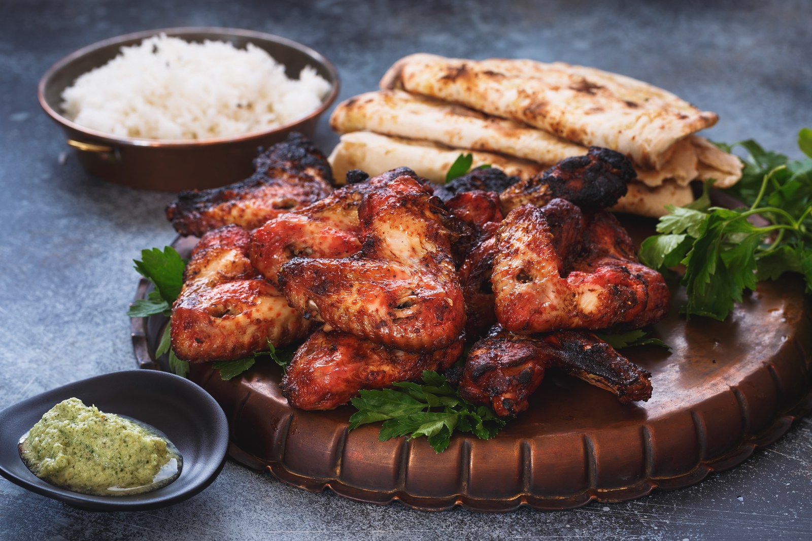 Charred tandoori chicken wings with deep red marinade and blackened edges piled on a wooden serving board with fresh cilantro, served with grilled naan bread, white basmati rice in a copper bowl, and green chutney