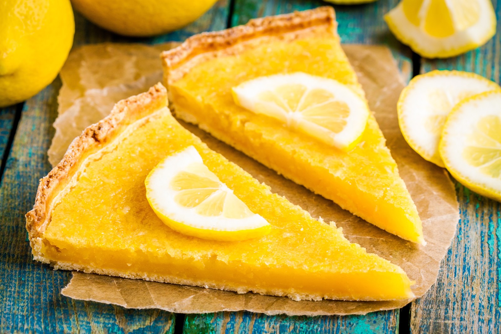 Two slices of French lemon tart with vibrant yellow lemon curd filling in a crisp shortcrust pastry shell, garnished with candied lemon slices, served on parchment paper with fresh lemons on a rustic turquoise wooden surface