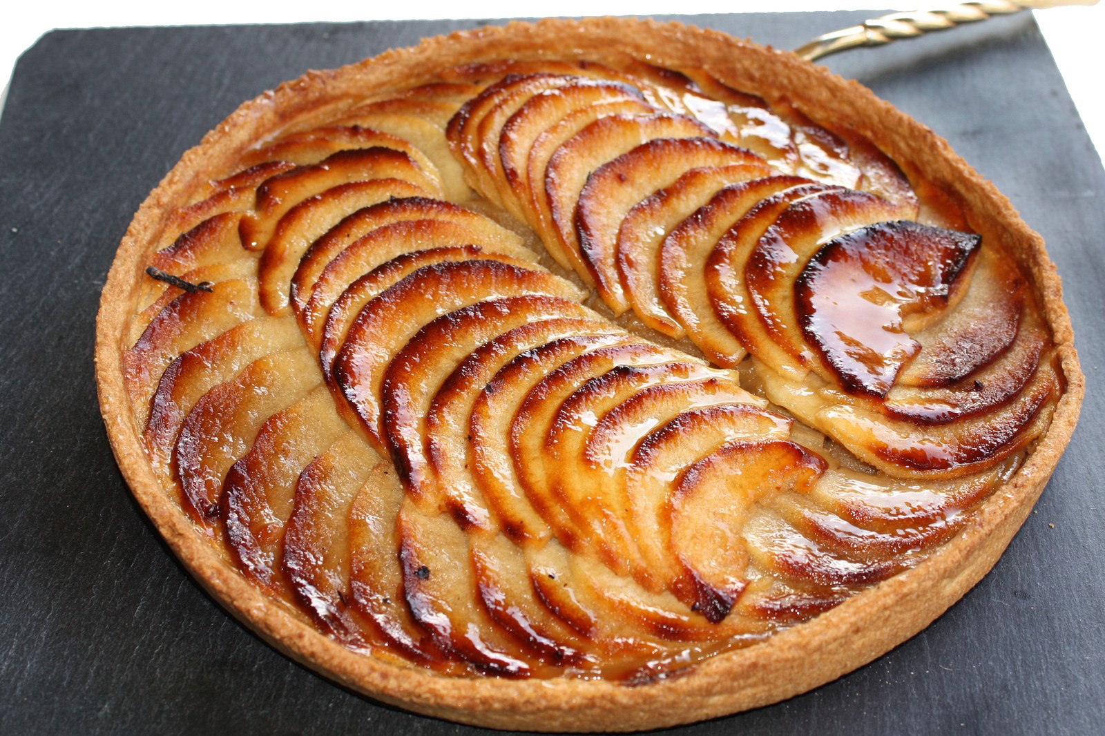 French apple tart with thin caramelized apple slices arranged in elegant spiral pattern, golden buttery crust, glossy apricot glaze on dark slate serving board