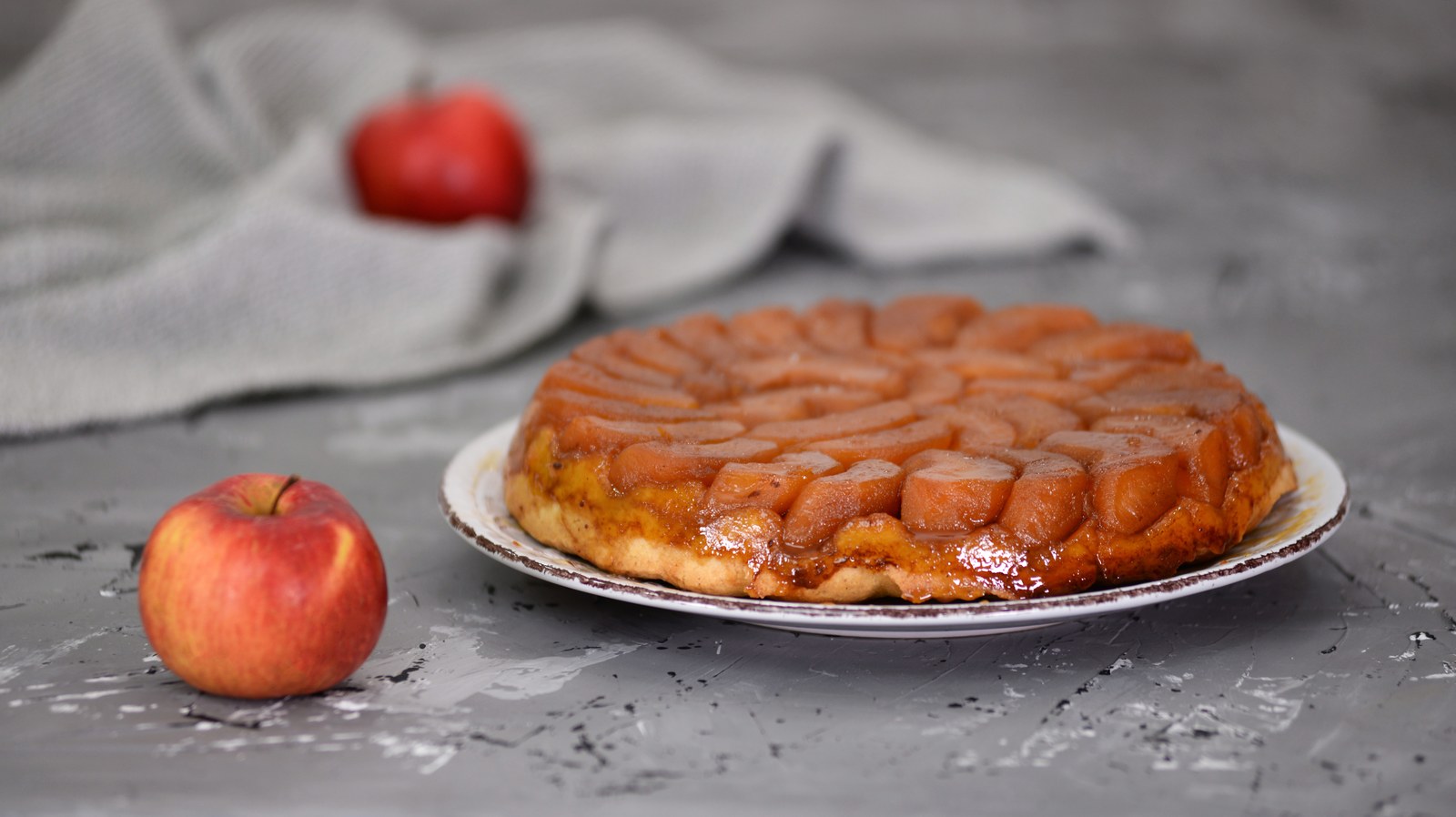 Classic French tarte tatin with caramelized apple halves arranged in a circular pattern, glazed with amber colored caramel sauce, baked upside down on buttery puff pastry crust, displayed on a white ceramic plate with fresh apples