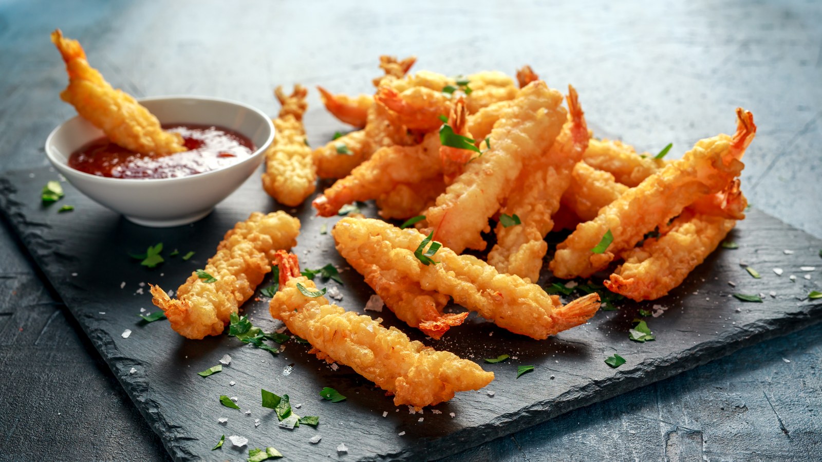 Japanese tempura shrimp with ultra-light crispy golden batter, fresh herbs, sweet chili dipping sauce in white bowl on black slate platter