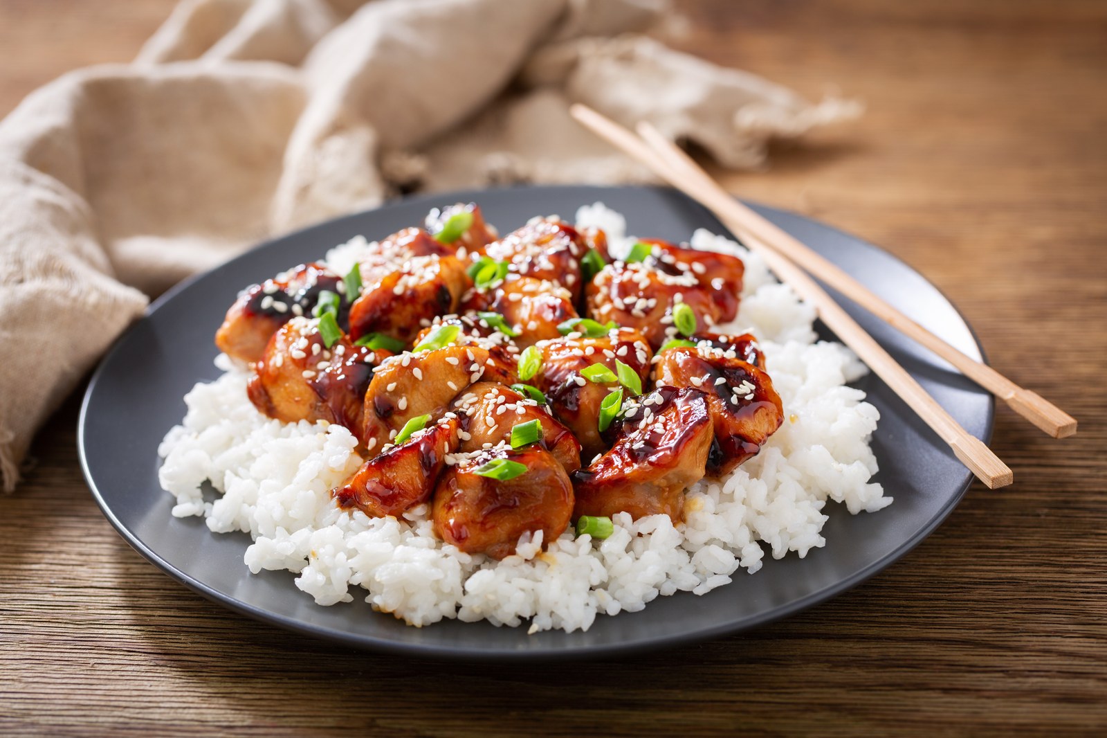 Teriyaki chicken pieces glazed in sweet soy sauce over steamed white rice, garnished with sesame seeds and sliced green onions on black plate