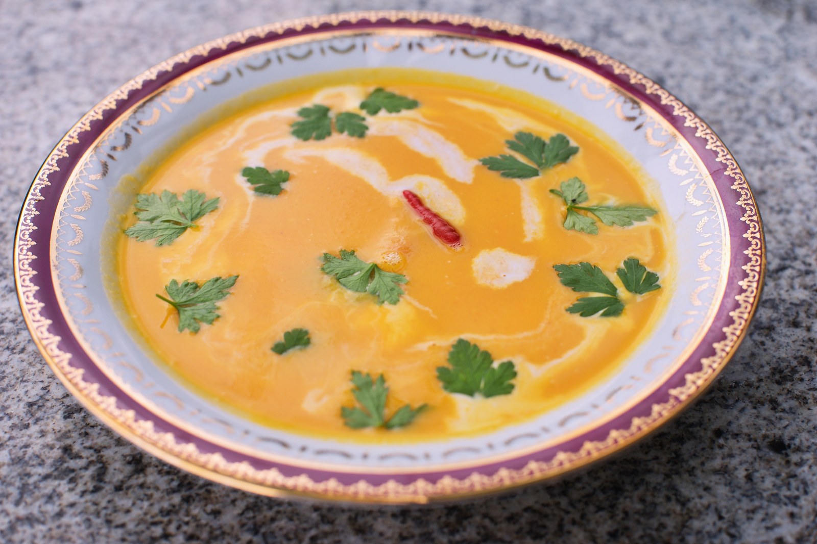 Creamy Thai coconut carrot soup in vibrant orange color, garnished with fresh cilantro, red chili, swirl of coconut cream in ornate purple-rimmed plate
