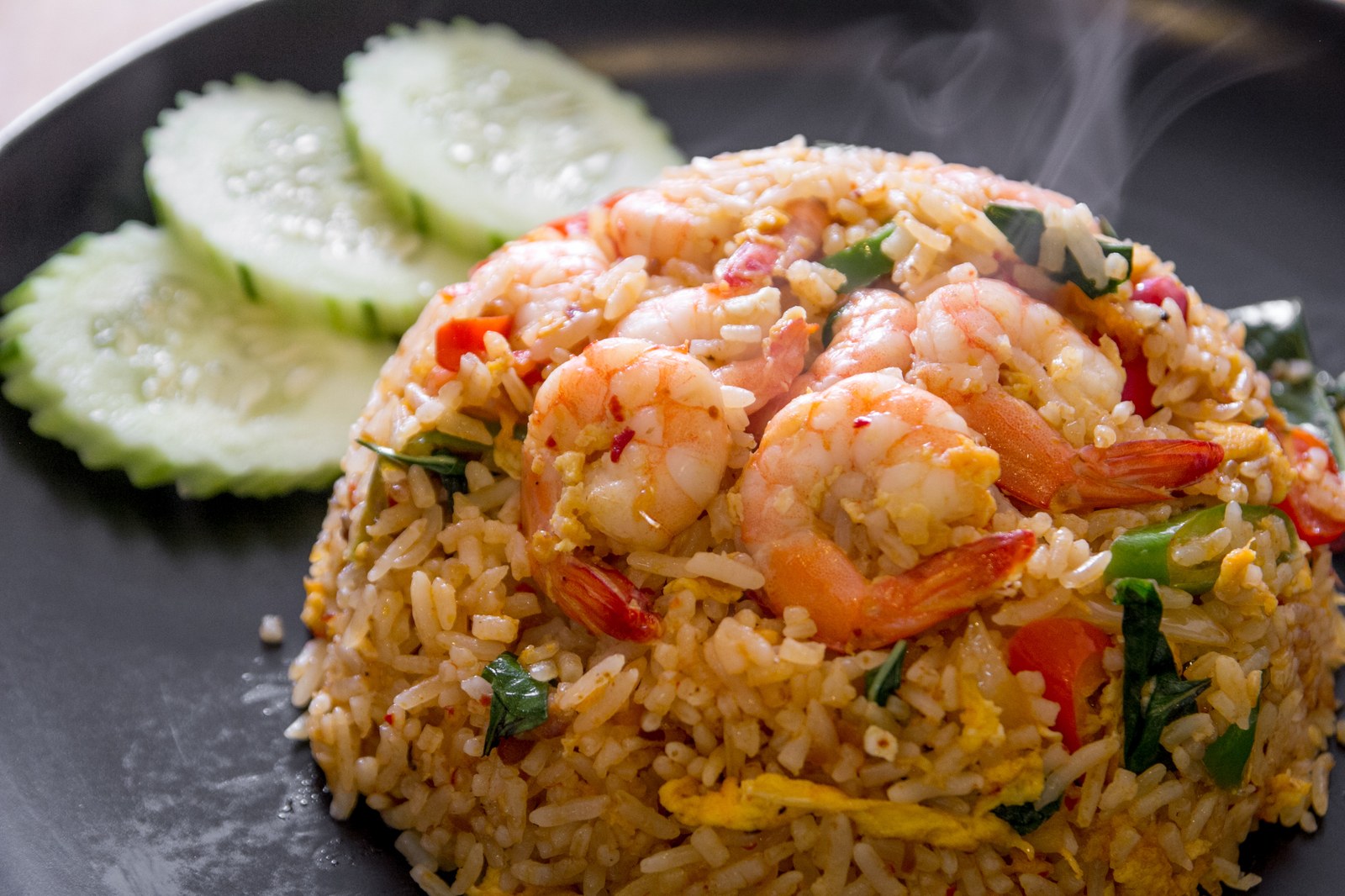 Thai fried rice with large shrimp, scrambled egg, Thai basil, red bell peppers, served with cucumber slices on black plate