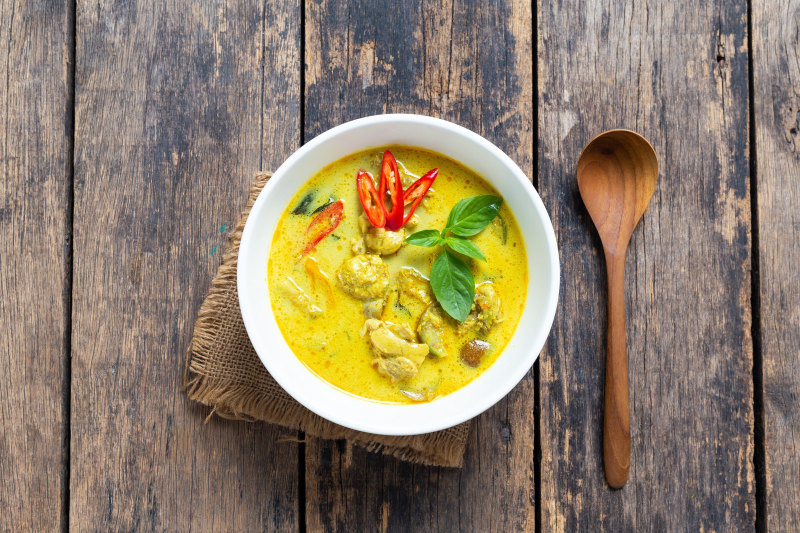 Thai green curry with chicken, bamboo shoots, Thai basil, red chilies in vibrant green coconut sauce in white bowl on burlap mat