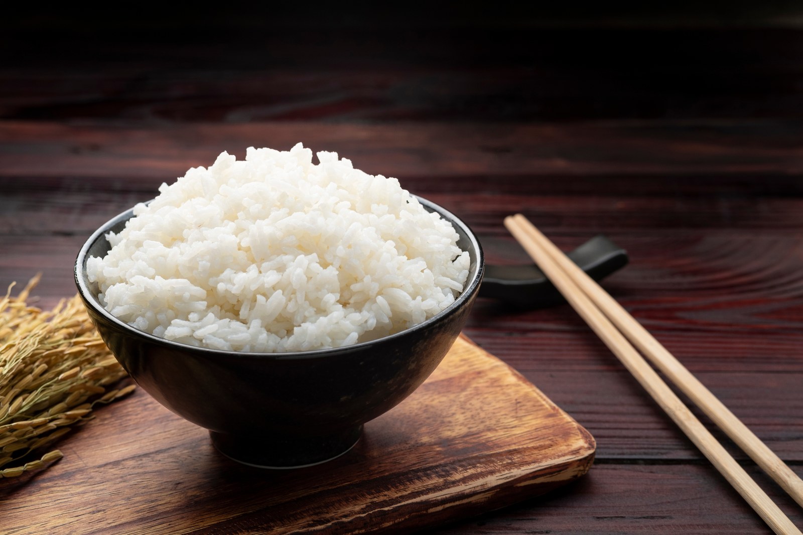 Bowl of steamed Thai sticky rice with glossy white grains in black ceramic bowl with raw rice stalks on dark wooden board