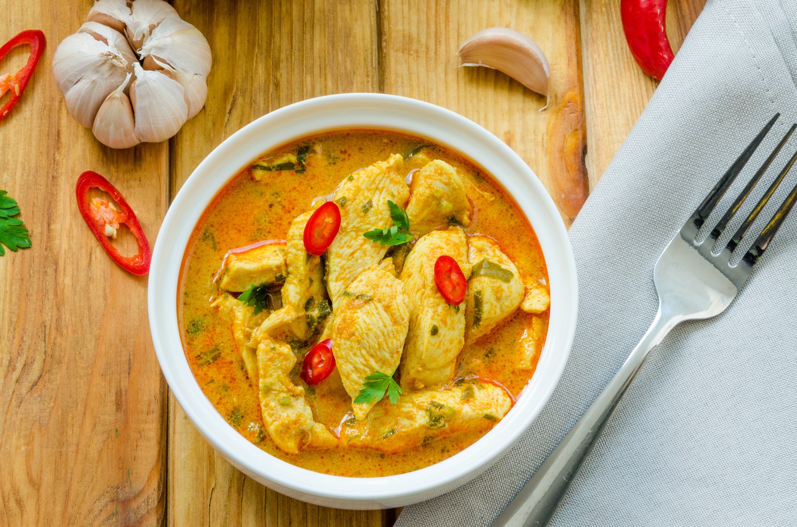 Thai yellow curry with chicken pieces in golden turmeric-coconut sauce, garnished with cilantro and red chilies in white bowl with wooden spoon