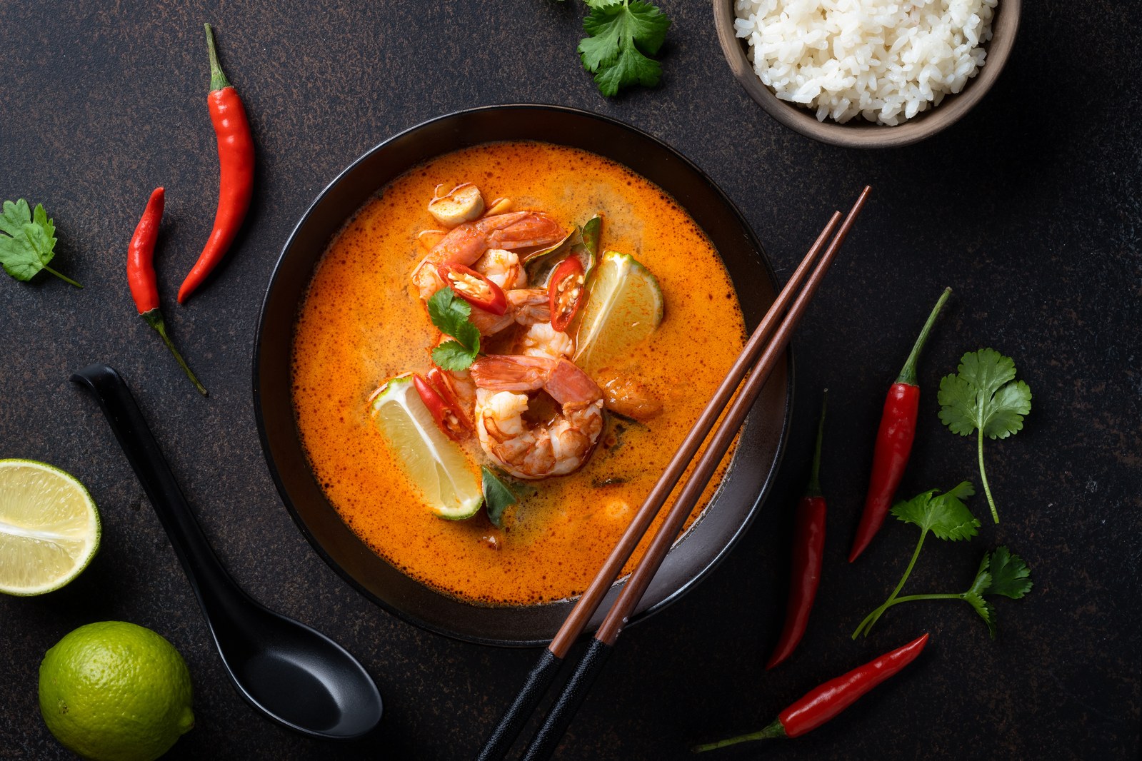 Thai tom yum soup with shrimp, lemongrass, lime wedges, red chilies in spicy orange-red broth in black bowl with chopsticks