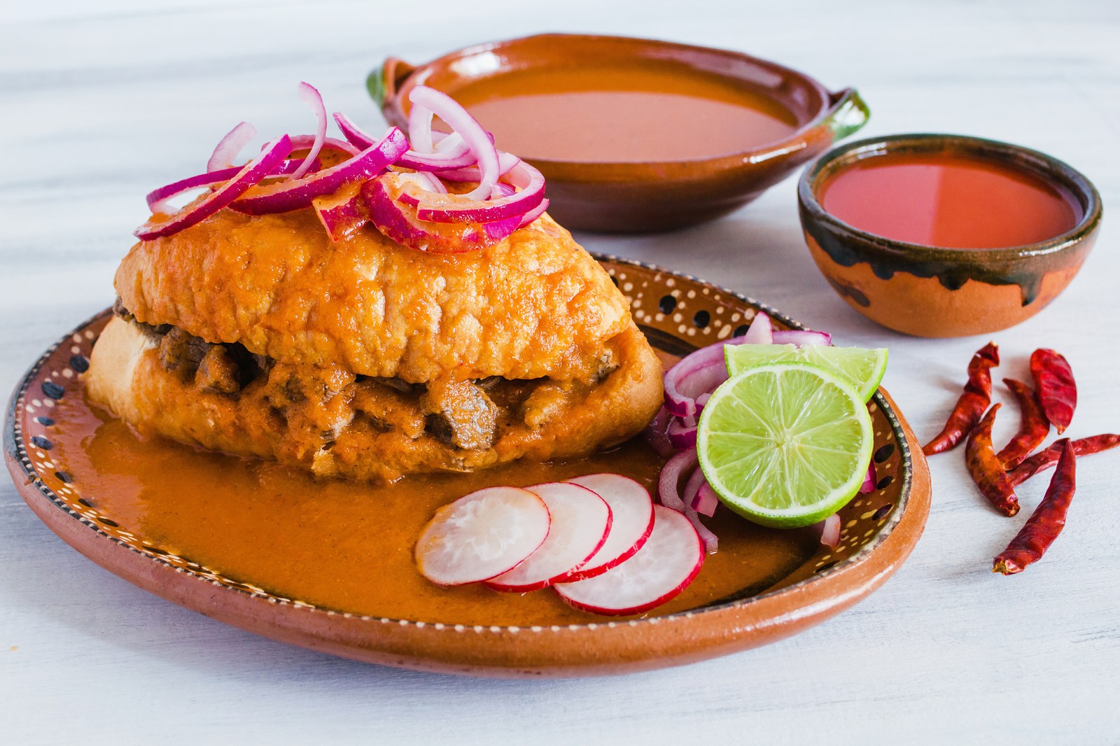 Mexican torta ahogada sandwich drowned in spicy red tomato sauce, topped with pickled red onions, served with lime wedges and radish slices on clay plate