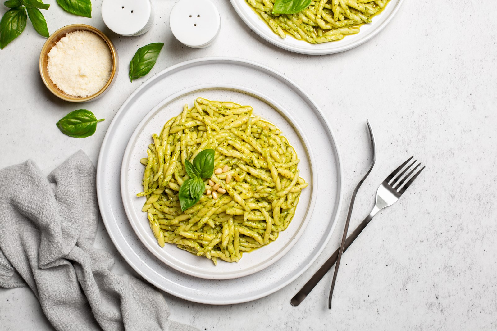 Italian trofie pasta in vibrant green Genovese basil pesto with toasted pine nuts, grated Parmesan, fresh basil leaves on white plates