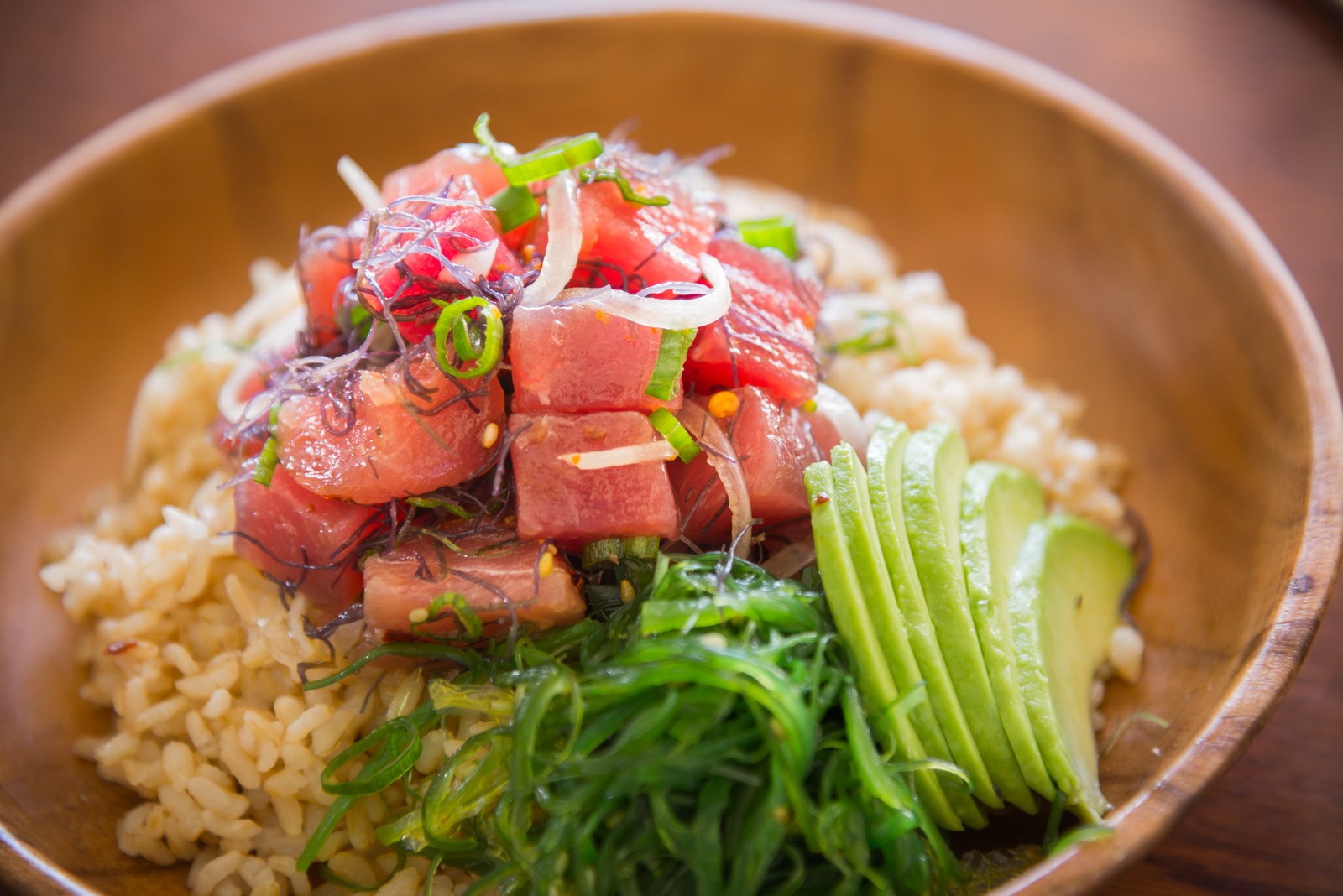 Hawaiian tuna poke bowl with ruby-red ahi tuna cubes, wakame seaweed salad, sliced avocado, scallions, sesame seeds over white rice in wooden bowl