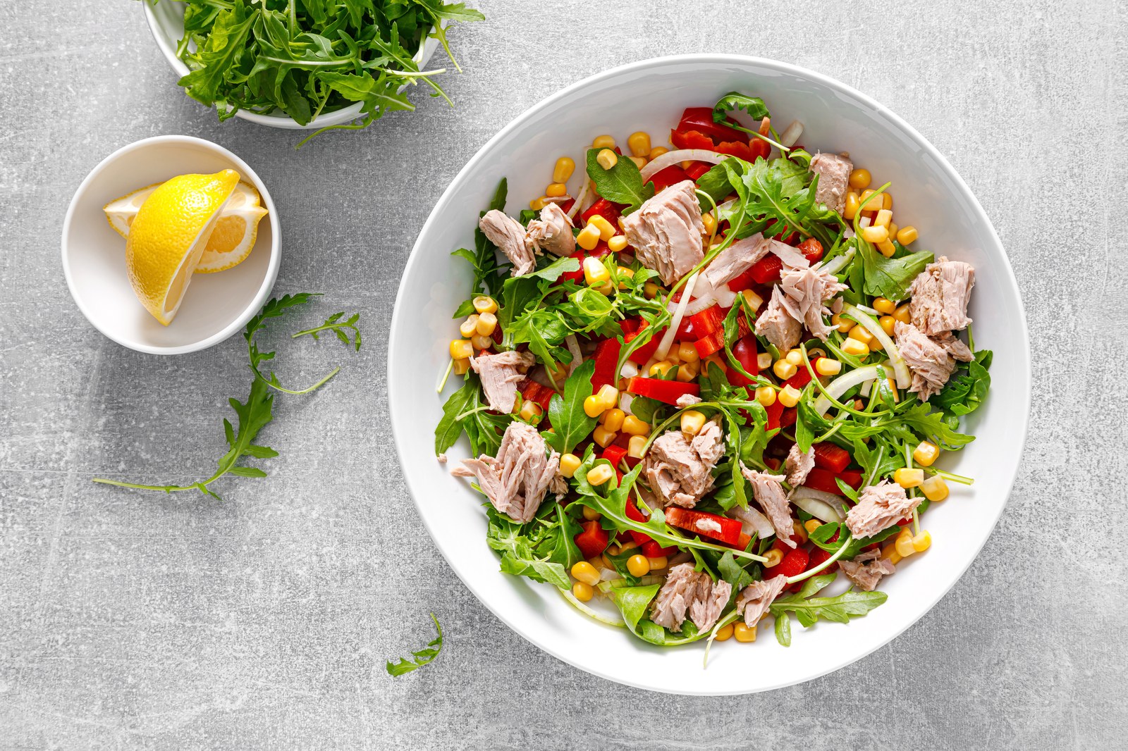 Fresh tuna salad with chunks of seared tuna, arugula, sweet corn kernels, red bell pepper slices, mixed greens in white bowl with lemon wedge