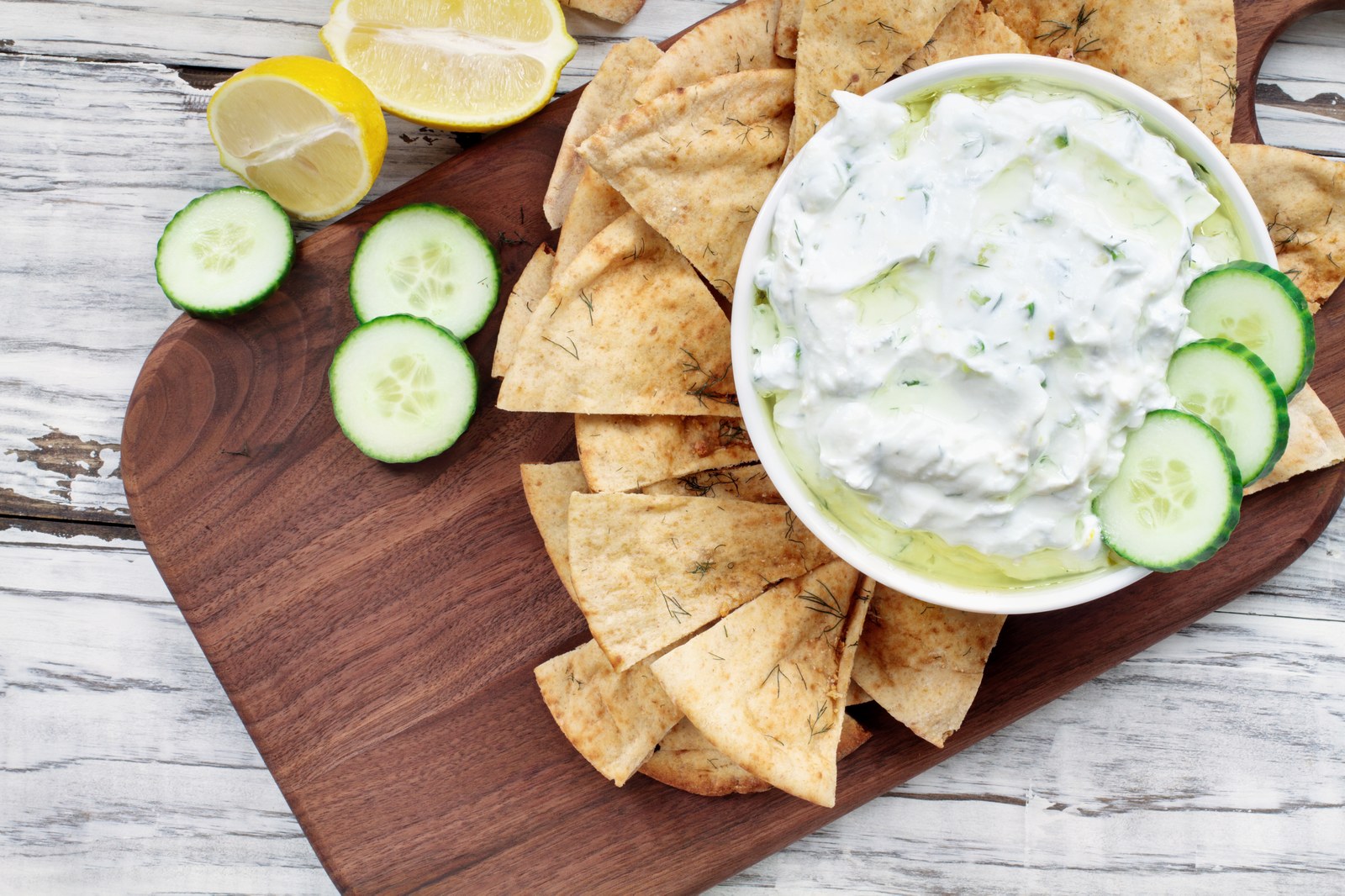 Greek tzatziki sauce with creamy yogurt, grated cucumber, fresh dill in white bowl, drizzled with olive oil, garnished with cucumber slices, served with herbed pita chips on wooden board