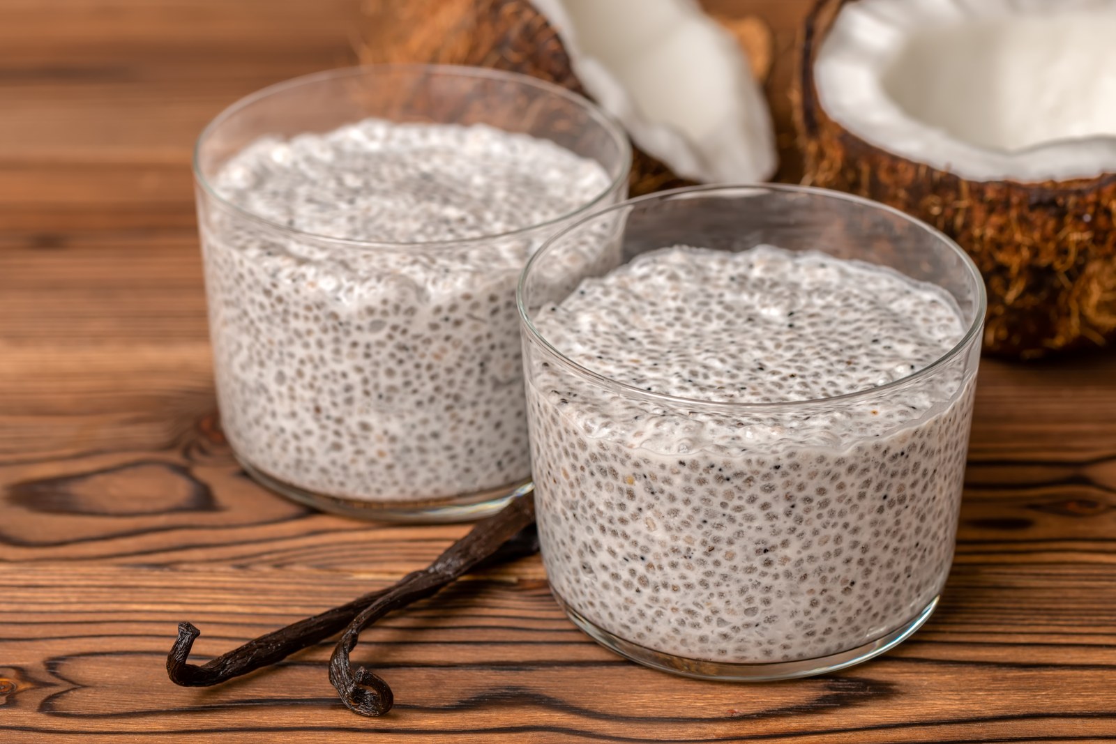 Vanilla bean chia seed pudding with swollen chia seeds in creamy coconut milk, whole vanilla beans, coconut halves on wooden board in clear glasses