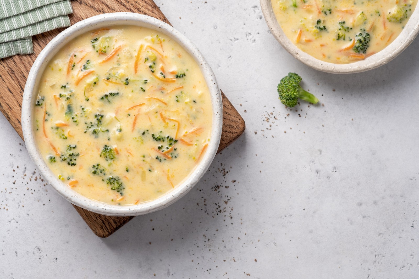 Creamy broccoli cheddar cheese soup with green broccoli florets, shredded orange cheddar, pale yellow cream base in white rustic bowls